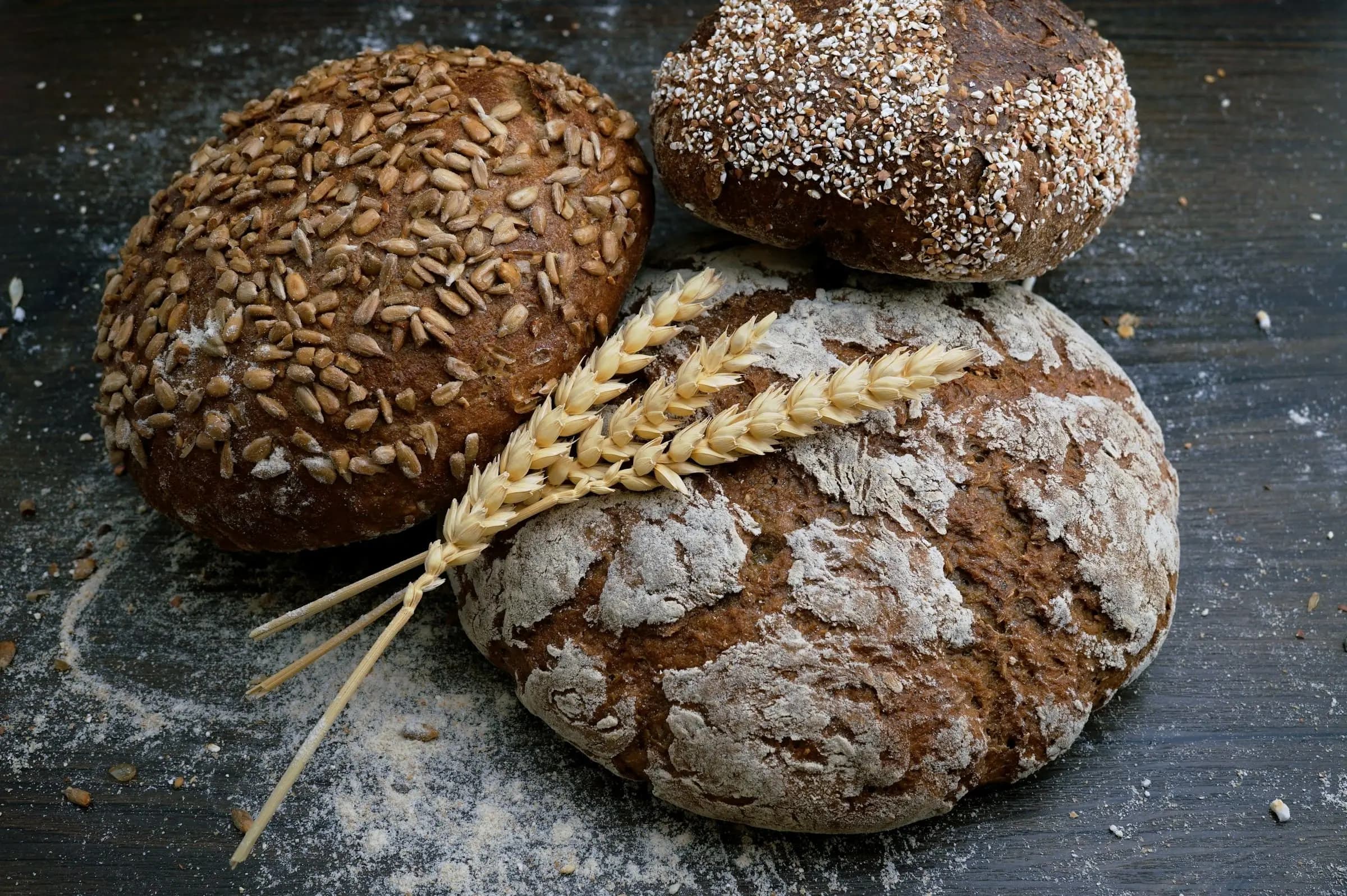 Three rustic whole-grain bread loaves topped with seeds beside wheat stalks on a dark wooden table